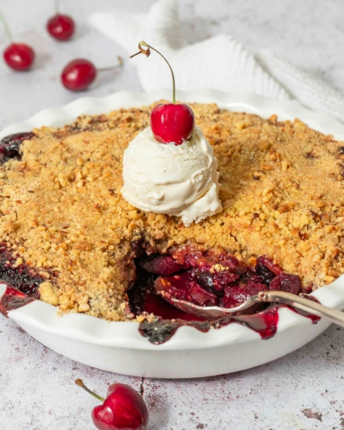 A freshly baked apple cherry crumble with vegan vanilla ice cream and a fresh red cherry on top