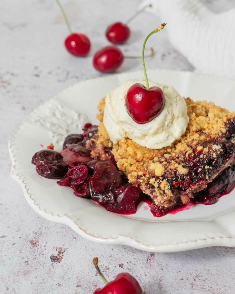 A serving plate of warm apple cherry crumble, with a golden hazelnut crumb topping, vanilla ice cream and a fresh red cherry on top