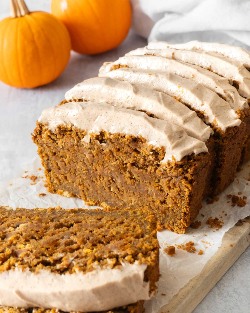 A sliced and frosted vegan gluten free pumpkin bread cake on a table top surrounded by mini pumpkins