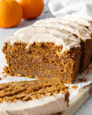 A sliced and frosted vegan gluten free pumpkin bread cake on a table top surrounded by mini pumpkins