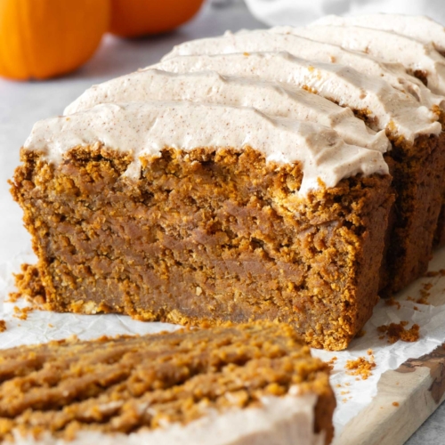 A sliced and frosted vegan gluten free pumpkin bread cake on a table top surrounded by mini pumpkins