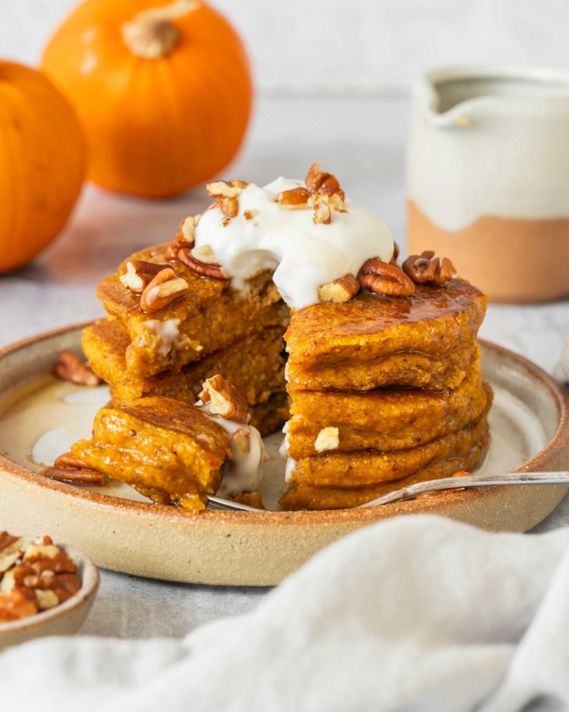 Vegan pumpkin pancakes topped with coconut yoghurt, crumbled pecans and maple syrup, with a slice cut out
