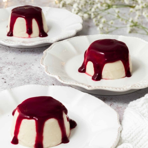 3 vegan panna cotta on white plates with raspberry coulis drizzled over them