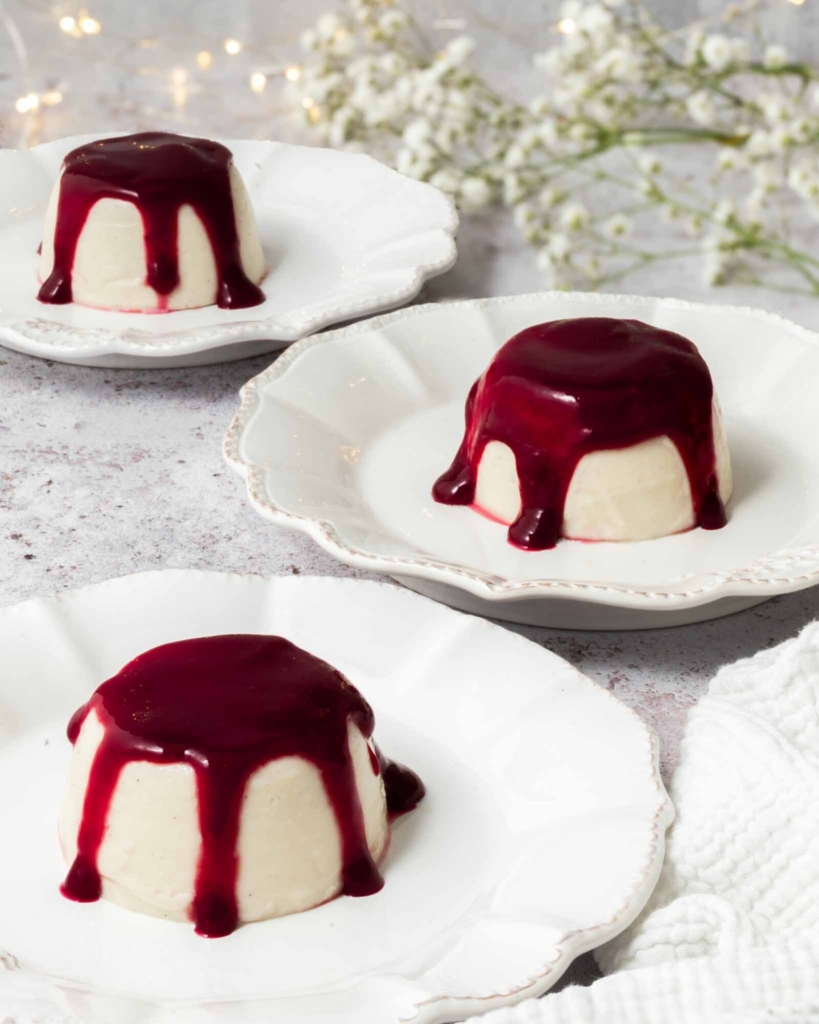 3 vegan panna cotta on white plates with raspberry coulis drizzled over them