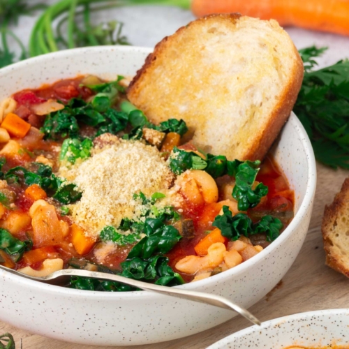 A close up image of a bowl of hearty vegetable and bean soup, topped with fresh cavolo nero and dairy free parmesan cheese