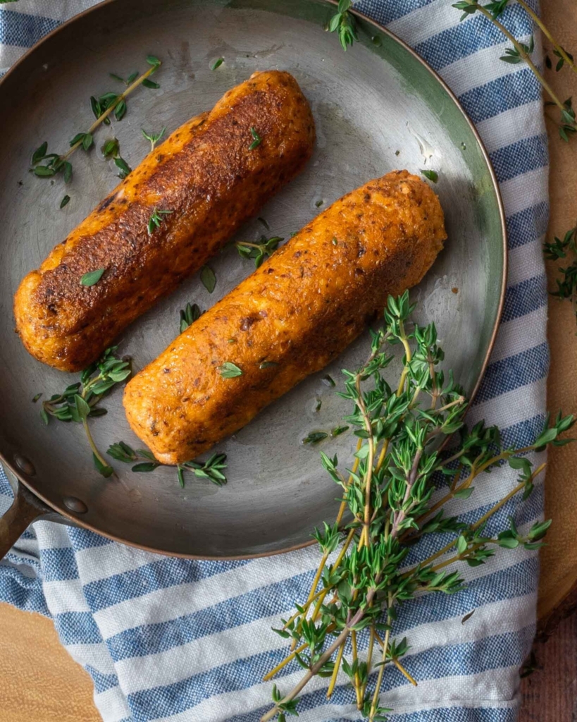 Two tofu sausages in a small frying pan, sprinkled in fresh herbs