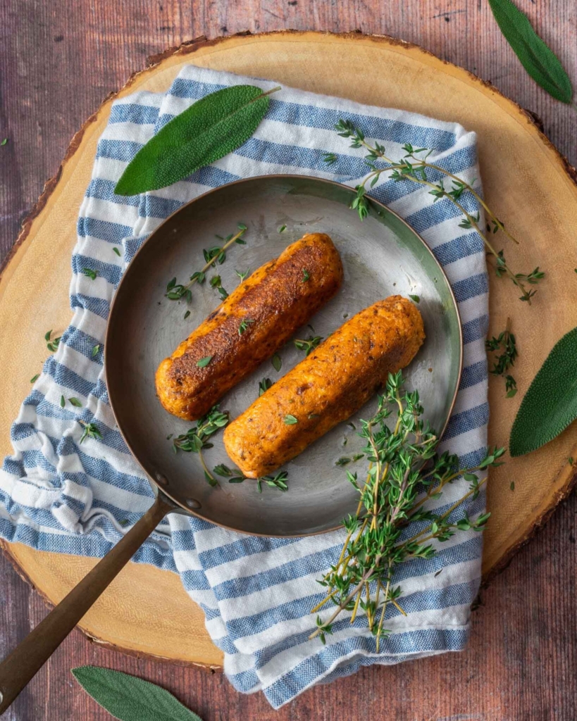 Two tofu sausages in a small frying pan, sprinkled in fresh herbs