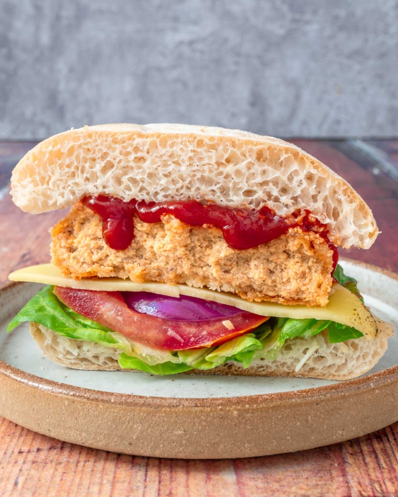 A homemade gluten free vegan chicken burger cut in half in a bun with ketchup drizzling down the side